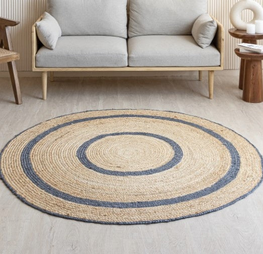 Round jute rug with blue border on a light wooden floor, with a gray sofa and wooden side table in the background.