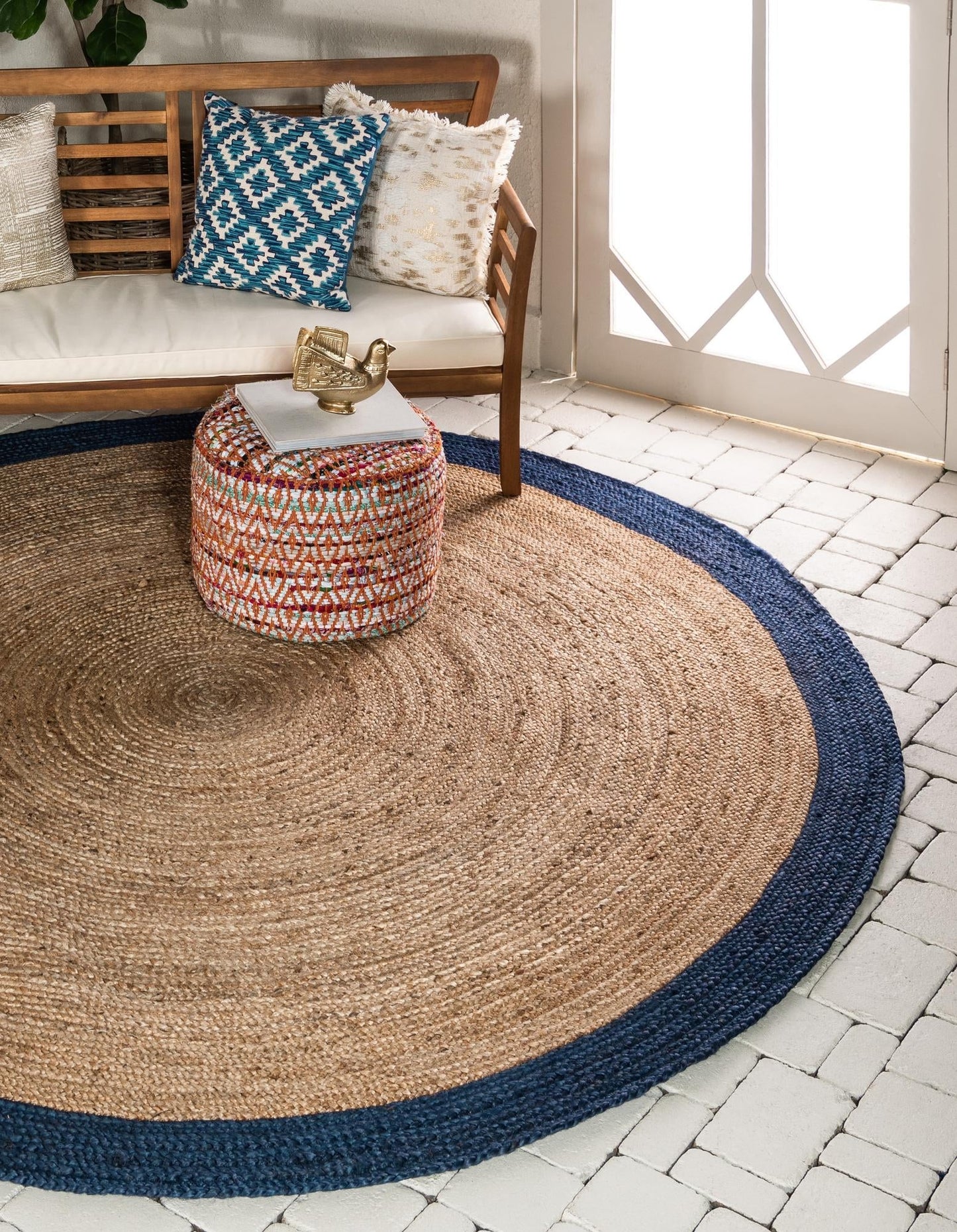 Round jute rug with blue border on a patio with a wooden bench and colorful ottoman.